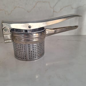 May include: A stainless steel potato ricer with a perforated basket and long handles. The kitchen utensil is designed for mashing potatoes and other soft foods. The ricer is made of shiny metal and has a classic design.