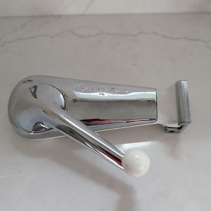 May include: A vintage chrome-plated can opener with a white handle and a white knob. The can opener has the text "Can-O-Mat" on the side. The can opener is lying on a white marble surface.