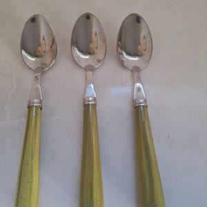 May include: Three spoons with oval-shaped stainless steel bowls and olive green handles. The handles have a tapered design and are connected to the spoon bowls with a silver band. The spoons are arranged side by side.