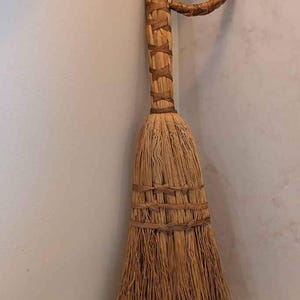 May include: A hand-held broom constructed from natural tan fibers. The handle features a circular loop at the top, wrapped in woven material. The bristles are bound together with a woven band, creating a flared shape at the base of the broom.