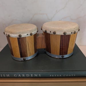May include: A set of two bongo drums featuring alternating vertical panels of light and dark wood. The drums have off-white drumheads secured with metal rims and studs, connected by a central wooden block, resting on two stacked books.