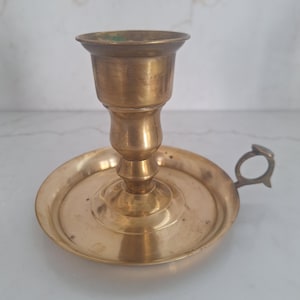 May include: A vintage brass candlestick holder with a round base, a central stem, and a cup-shaped top. It features a small handle on the side. The brass has a warm, aged patina, suggesting its antique nature. The candlestick holder is designed to hold a single candle.