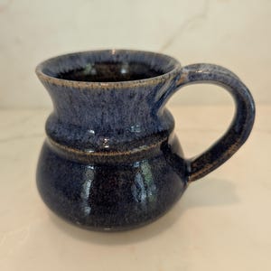 May include: A ceramic mug with a rounded, bulbous base and a flared rim. The mug features a deep blue glaze with mottled, lighter blue and grey tones throughout the surface. It includes a single curved handle attached to the side of the body.