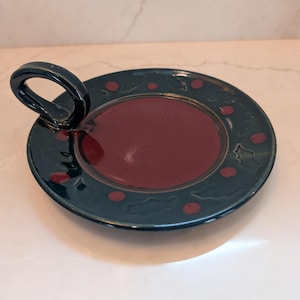 May include: A round ceramic candle holder with a dark green rim featuring embossed holly leaf patterns and red berry accents. The center of the plate is a deep, glossy maroon color. A single, curved, dark green handle is attached to the side.