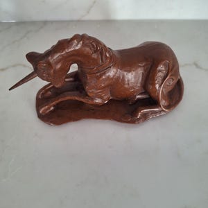 May include: A brown, detailed sculpture of a unicorn in a resting position. The mythical creature has a long horn and is lying on a textured base. The sculpture is made of a dark brown material and is set against a white background.