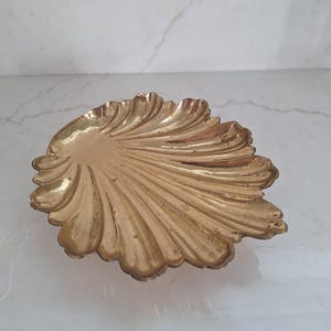 May include: A gold-colored, seashell-shaped decorative dish. The dish has a detailed, ridged surface, with a scalloped edge. The dish is made of metal and is likely used for decorative purposes or to hold small items.