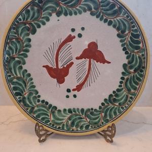 May include: A round, hand-painted ceramic plate with a white center featuring two red bird-like figures. The rim is decorated with green leaves and a yellow border. The plate is displayed on a decorative metal stand.