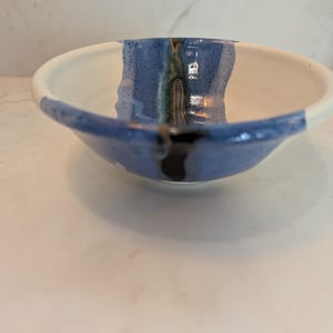 May include: A ceramic bowl with a smooth white interior and a wide, textured blue band running vertically down the center. The blue section features a dark, glossy drip pattern at the rim and base, with subtle green and brown undertones.