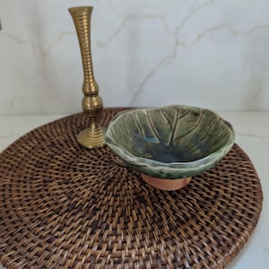 May include: A tall, slender brass candlestick with a ribbed stem and a wide circular base, positioned next to a small, shallow ceramic bowl shaped like a green leaf with visible veins and a smooth, glossy finish on a round woven mat.