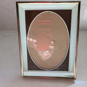 May include: A rectangular picture frame with a gold trim and a light green border. The frame has an oval opening with a brown backing and a clear glass cover. The frame is designed to hold a photograph or artwork.