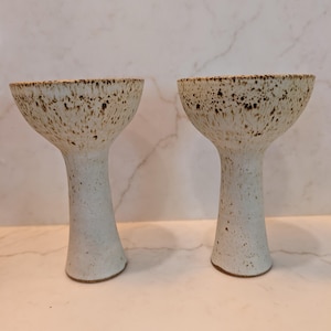 May include: Two ceramic goblets featuring a speckled off-white glaze. Each vessel has a wide, flared bowl that tapers down to a slender, cylindrical stem and a circular base. The upper rims are heavily textured with dark brown speckles.