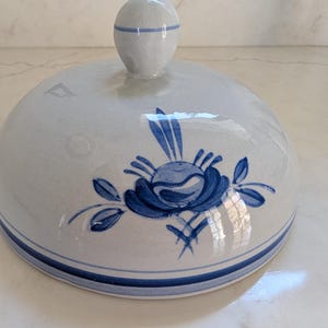 May include: A ceramic lid with a blue floral design. The lid is light blue with a blue floral pattern and a blue stripe around the edge. A small, round handle is on top.