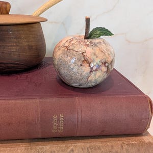 May include: A polished stone apple figurine with mottled pink, gray, and brown patterns, featuring a brown twig-like stem and a single green leaf, resting on a dark red book titled Empire Edition stacked atop a brown book.