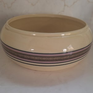 May include: A cream-colored ceramic bowl with a wide, rounded shape. The bowl features a decorative band of brown and pink stripes, with thin black lines above and below. The interior of the bowl is a light tan color.