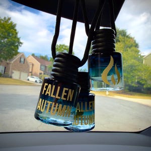 Op de afbeelding: Drie glazen auto-luchtverfrissers met zwarte touwhangers. De flessen hebben een blauwe en turquoise kleurovergang met gouden tekst "FALLEN AUTUMN". Eén fles heeft een gouden vlammenafbeelding.
