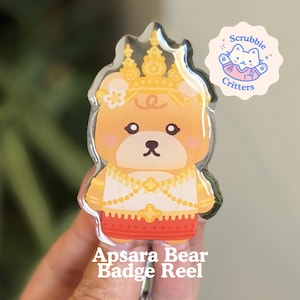 May include: A clear badge reel featuring a cartoon bear dressed in a gold crown and traditional Thai clothing. The bear has a cute expression and is labeled "Apsara Bear Badge Reel". A small logo with a cat is in the upper right corner.