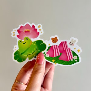 May include: Two stickers featuring cute illustrations. One shows a smiling green frog on a lily pad with a pink lotus flower. The other depicts pink triangular shapes with a bear and bunny.