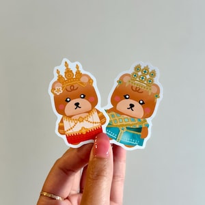 May include: Two stickers featuring cartoon bears in traditional Thai outfits. One bear wears a red and white outfit, the other a blue and gold outfit. Both bears have gold crowns and are smiling.