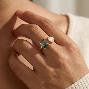 May include: A gold ring featuring two gemstones: a square, green stone and a pear-shaped, white stone. The ring is worn on a finger, showcasing the jewelry's design and color contrast. The ring's band is a thin, gold color.