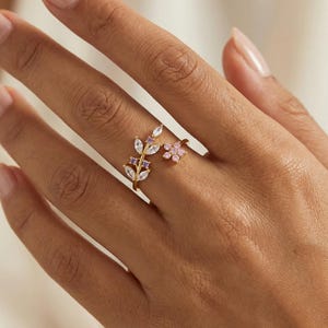Könnte beinhalten: Ein goldfarbener Ring mit floralem und Blätter-Design. Der Ring hat einen astähnlichen Aufbau mit klaren und violetten Edelsteinen sowie einer kleinen rosa Blume. Der Ring wird an einer Hand mit neutral gefärbten Fingernägeln getragen.