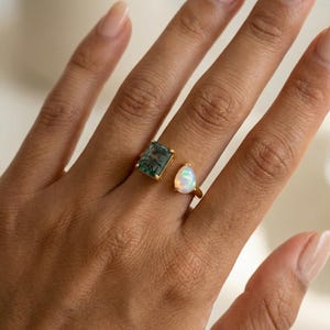 May include: Two gold rings with gemstones. One ring features a square, green gemstone, and the other has a pear-shaped, iridescent opal. The rings are displayed on a hand.