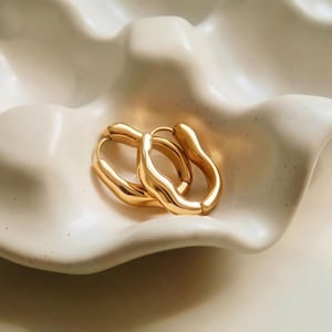 May include: A pair of gold-colored hoop earrings with an organic, wavy design. The earrings are displayed on a white, textured surface, creating a minimalist and elegant aesthetic. The earrings have a polished finish, reflecting light.