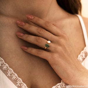 May include: A gold ring featuring two gemstones: a rectangular green stone and a smaller, round white stone. The ring is worn on a finger, with the gemstones positioned on either side of the finger.