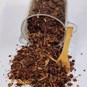 May include: A clear glass jar, tilted to spill brown dried herbs and bark onto a white surface. A wooden spoon lies beside the spill. The contents appear to be a natural product, possibly for herbal remedies.