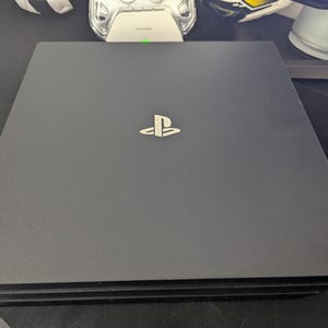 May include: A matte black rectangular gaming console featuring a centered silver PlayStation logo on the top surface. The device has a layered, tiered design with a slim profile and a smooth, flat finish across the main top panel.