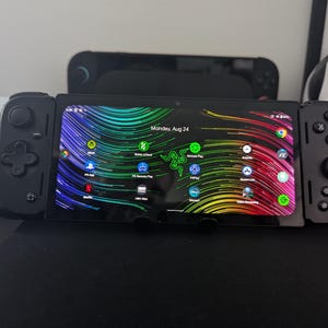May include: A black handheld gaming device with a vibrant, multi-colored abstract wallpaper on the screen. The device features integrated controllers on both sides with a D-pad, analog sticks, and standard action buttons. The screen displays app icons.