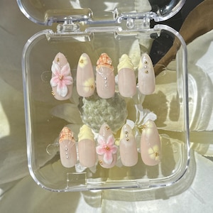 May include: A set of ten artificial nails in a clear plastic case. The nails are a light pink color with various designs, including 3D flowers, yellow tips, and gold accents. The nails are oval-shaped and appear to be ready for application.