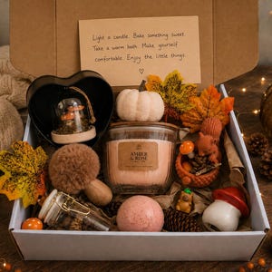 May include: A gift box containing a candle labeled Amber & Rose, a white pumpkin, a glass jar with mushrooms, a pink bath bomb, a squirrel figurine, a red mushroom ornament, faux autumn leaves, and a note reading Light a candle. Bake something sweet.