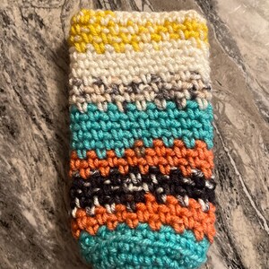 May include: A handmade crochet cozy in a rectangular shape. The cozy features horizontal stripes of yellow, white, teal, orange, gray, and black. The item is likely designed to hold a beverage container.