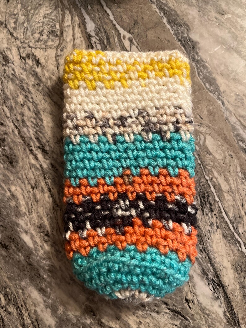 May include: A handmade crochet cozy in a rectangular shape. The cozy features horizontal stripes of yellow, white, teal, orange, gray, and black. The item is likely designed to hold a beverage container.