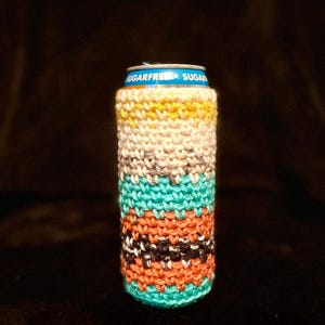 May include: A colorful, hand-knitted can cozy. The cozy features horizontal stripes in various colors, including yellow, cream, teal, orange, black, and white. The top of the can is visible, with the words "SUGARFREE" printed on it.