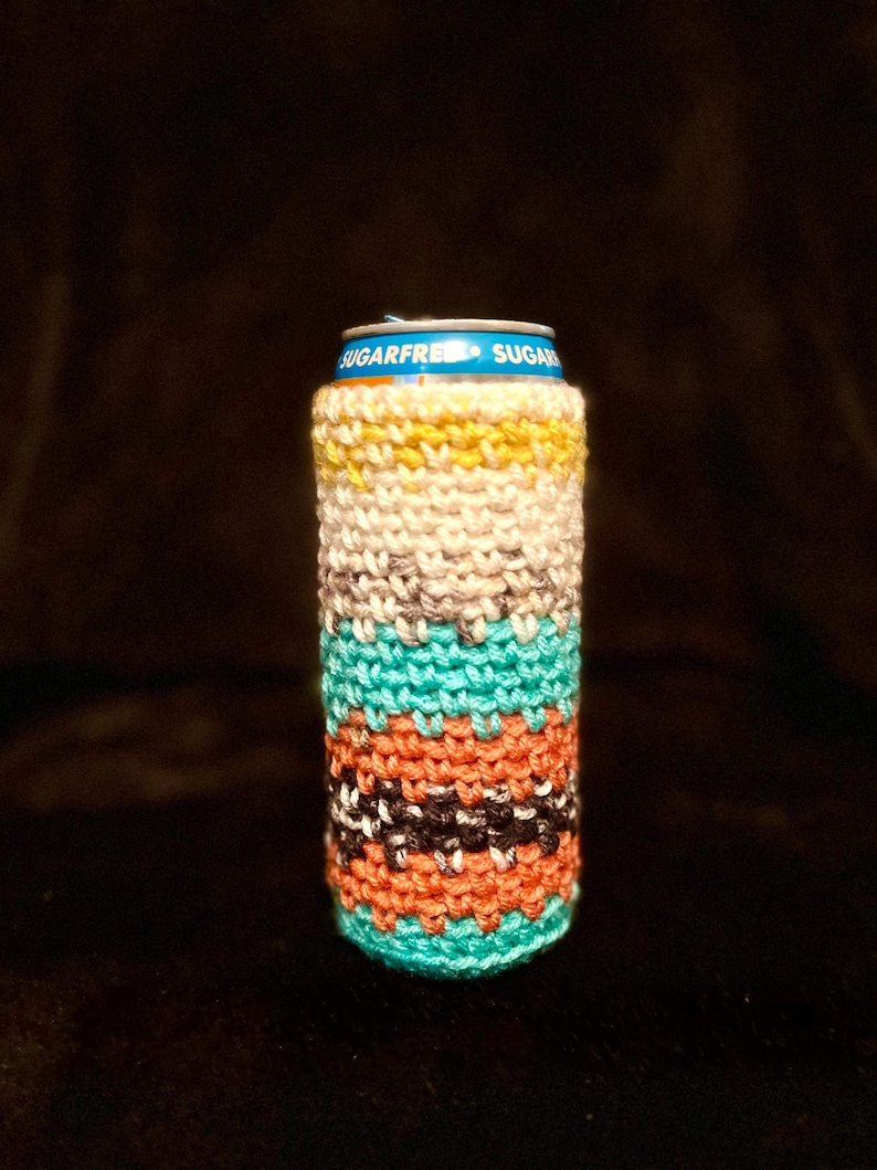May include: A colorful, hand-knitted can cozy. The cozy features horizontal stripes in various colors, including yellow, cream, teal, orange, black, and white. The top of the can is visible, with the words "SUGARFREE" printed on it.