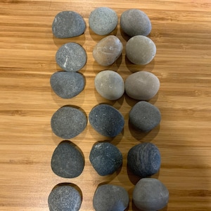 May include: A collection of smooth, gray river rocks of various sizes and shapes. The stones are arranged in neat rows on a light brown wooden surface. The rocks vary in color from light to dark gray.