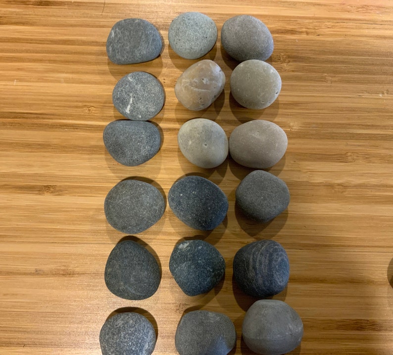 May include: A collection of smooth, gray river rocks of various sizes and shapes. The stones are arranged in neat rows on a light brown wooden surface. The rocks vary in color from light to dark gray.