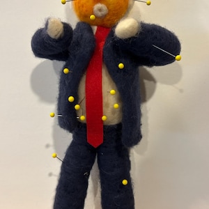 May include: A needle-felted doll with orange skin, yellow hair, and a white shirt, wearing a dark blue suit jacket and trousers with a bright red tie. The figure is pierced with multiple yellow-headed sewing pins across the face and body.