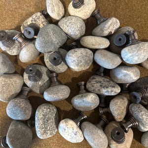 May include: A collection of gray stone cabinet knobs with dark bronze-colored metal hardware. The knobs are made from natural river stones, each with a unique shape and texture. The hardware includes a threaded post for easy installation.