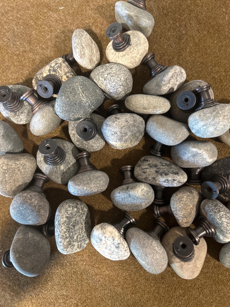 May include: A collection of gray stone cabinet knobs with dark bronze-colored metal hardware. The knobs are made from natural river stones, each with a unique shape and texture. The hardware includes a threaded post for easy installation.