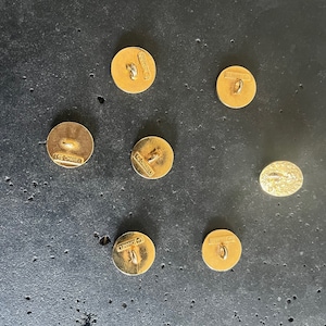 May include: A collection of seven gold-toned buttons, including one oval-shaped button. The round buttons have a loop on the back for sewing and the word "CHANEL" is stamped on the back of each button. The buttons are on a dark gray surface.