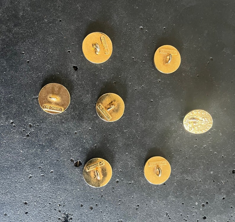 May include: A collection of seven gold-toned buttons, including one oval-shaped button. The round buttons have a loop on the back for sewing and the word "CHANEL" is stamped on the back of each button. The buttons are on a dark gray surface.