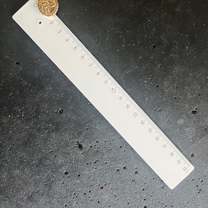 May include: A gold-colored coin with the year 1999 visible, placed near the top of a white ruler. The ruler is marked with black numbers and lines, measuring up to 20 inches. The coin and ruler rest on a dark, textured surface.