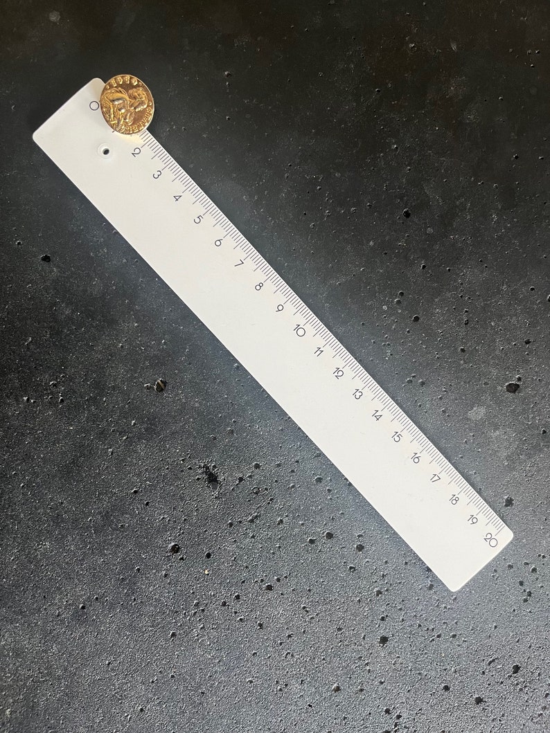 May include: A gold-colored coin with the year 1999 visible, placed near the top of a white ruler. The ruler is marked with black numbers and lines, measuring up to 20 inches. The coin and ruler rest on a dark, textured surface.