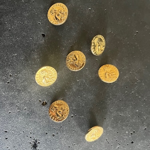 May include: Seven gold-colored buttons with embossed designs are scattered on a dark gray surface. Each button features a detailed relief, possibly a classical figure or emblem, and text around the edge. The buttons are circular and appear to be made of metal.