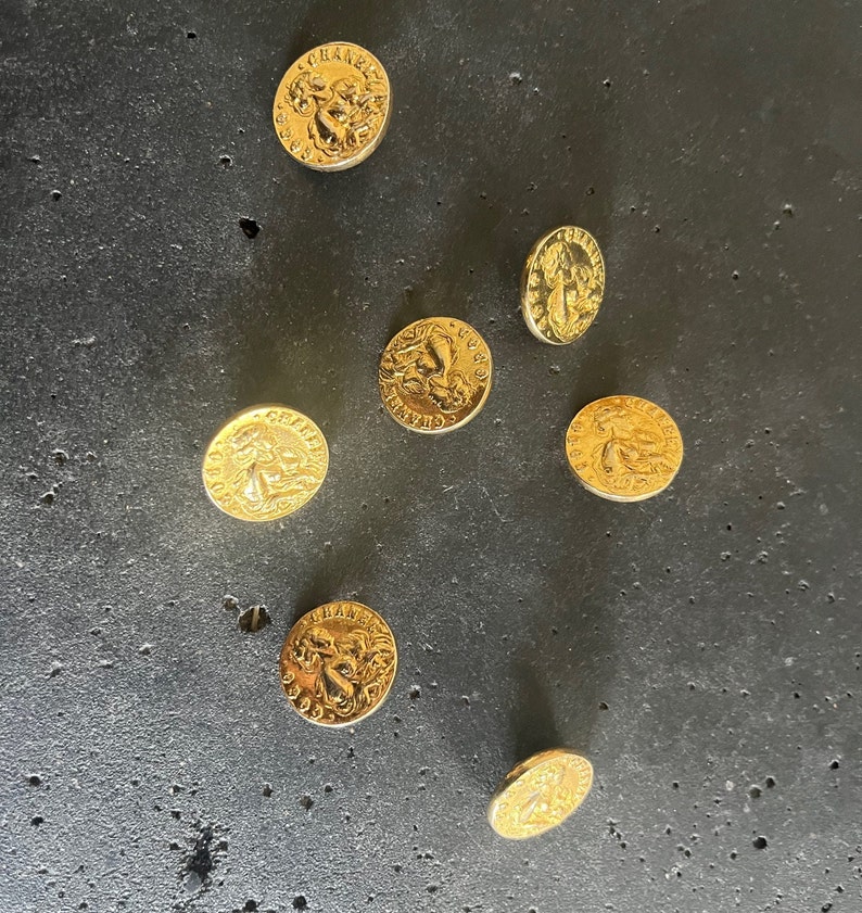 May include: Seven gold-colored buttons with embossed designs are scattered on a dark gray surface. Each button features a detailed relief, possibly a classical figure or emblem, and text around the edge. The buttons are circular and appear to be made of metal.