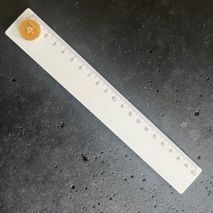 May include: A white ruler with black markings and numbers from 1 to 20 inches, with a gold-colored button on the left side. The ruler is lying on a dark gray surface.