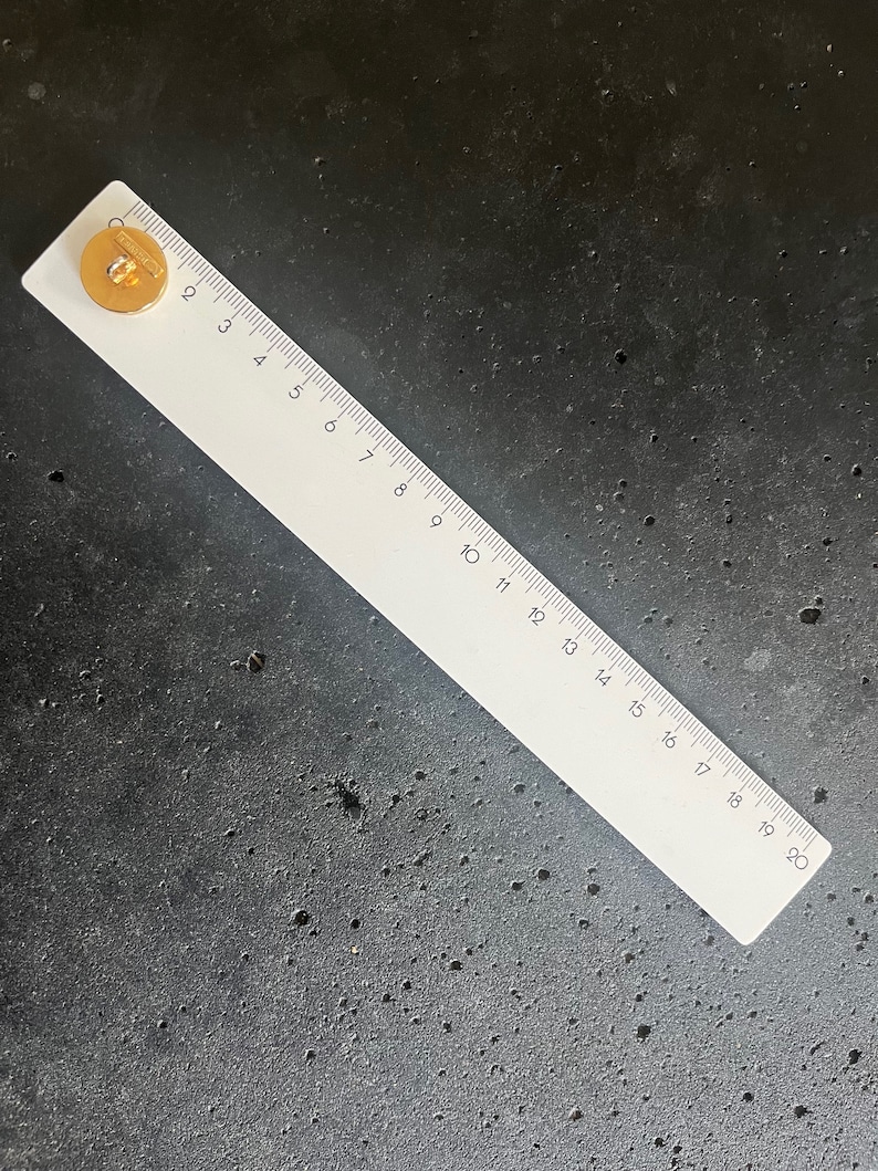 May include: A white ruler with black markings and numbers from 1 to 20 inches, with a gold-colored button on the left side. The ruler is lying on a dark gray surface.