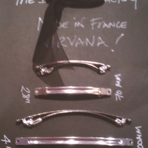 May include: Four silver hair clips of varying sizes. The clips are arranged vertically on a black surface. The text "The Factory" and "Made in France Nirvana!" is written in white on the surface.